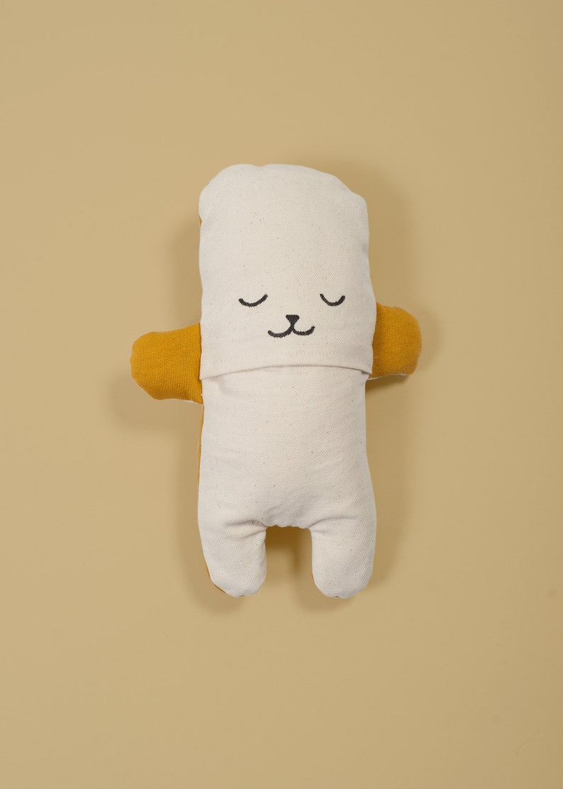 Pu&ograve; includere: Un giocattolo di peluche bianco con un viso e braccia in tessuto giallo. Il giocattolo ha un viso ricamato nero semplice con gli occhi chiusi e un piccolo naso.