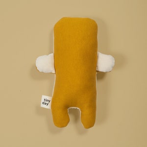 Pu&ograve; includere: Un giocattolo di peluche giallo senape con dettagli bianchi. Il giocattolo ha una forma semplice, umana, con braccia, gambe e una testa. Il giocattolo ha un piccolo cartellino che dice "tiny day".