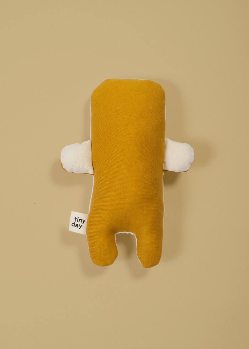 Pu&ograve; includere: Un giocattolo di peluche giallo senape con dettagli bianchi. Il giocattolo ha una forma semplice, umana, con braccia, gambe e una testa. Il giocattolo ha un piccolo cartellino che dice "tiny day".