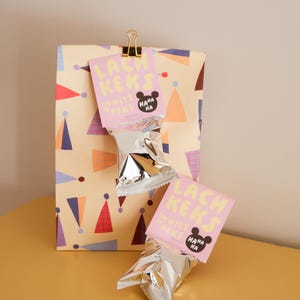 Könnte beinhalten: Eine Geschenkverpackung mit einem farbenfrohen geometrischen Muster, obenauf ein goldener Clip, der ein rosa Etikett mit der Aufschrift "LACH KEKS" und "UNWITZ KEKS" mit einem Cartoon-Bären hält. Zwei silberne Folienverpackungen sind befestigt.