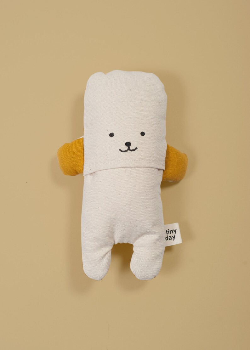 Pu&ograve; includere: Un giocattolo di peluche bianco con un semplice viso ricamato e braccia gialle. Il giocattolo ha un'etichetta che dice "tiny day".