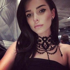 May include: A woman with long black hair wearing a black choker necklace with a beaded fringe. The necklace is made of black lace and beads.