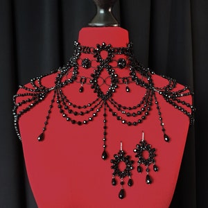 May include: A black beaded choker necklace and matching shoulder piece with dangling earrings. The jewelry is made of black beads and has a delicate, intricate design.