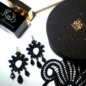 May include: A pair of black beaded earrings with a circular design and dangling teardrop beads. The earrings are displayed on a white surface with a black lace fabric, a black perfume bottle with gold accents, and a black round box with gold accents.