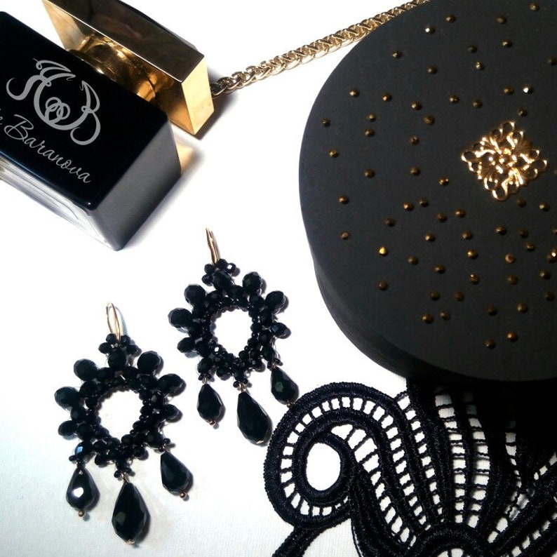 May include: A pair of black beaded earrings with a circular design and dangling teardrop beads. The earrings are displayed on a white surface with a black lace fabric, a black perfume bottle with gold accents, and a black round box with gold accents.