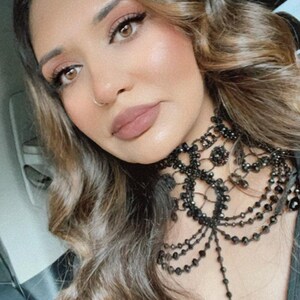 May include: A woman with long brown hair wearing a black beaded choker necklace with a lace design. The necklace has a long, dangling pendant.