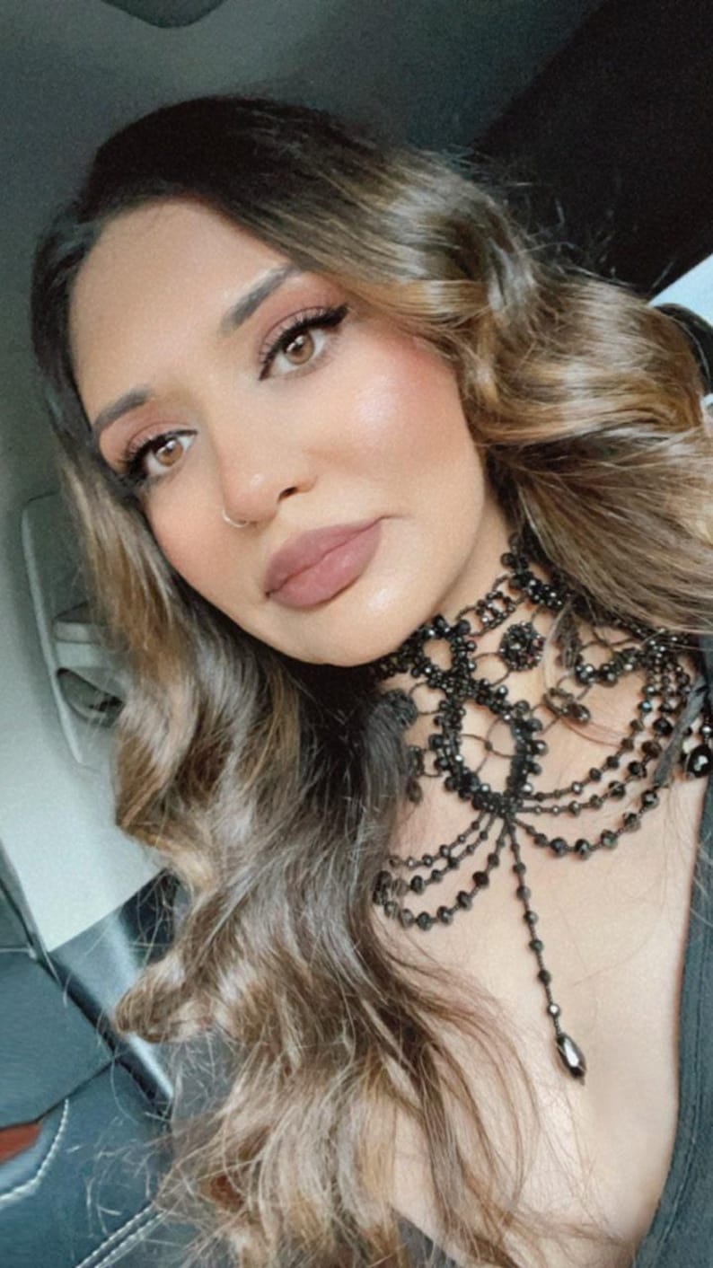 May include: A woman with long brown hair wearing a black beaded choker necklace with a lace design. The necklace has a long, dangling pendant.