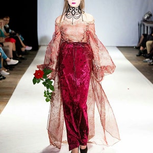 May include: A model walks the runway wearing a long, red velvet skirt with a sheer, gold-patterned overlay. The skirt is paired with a sheer, off-the-shoulder top and a black beaded choker.