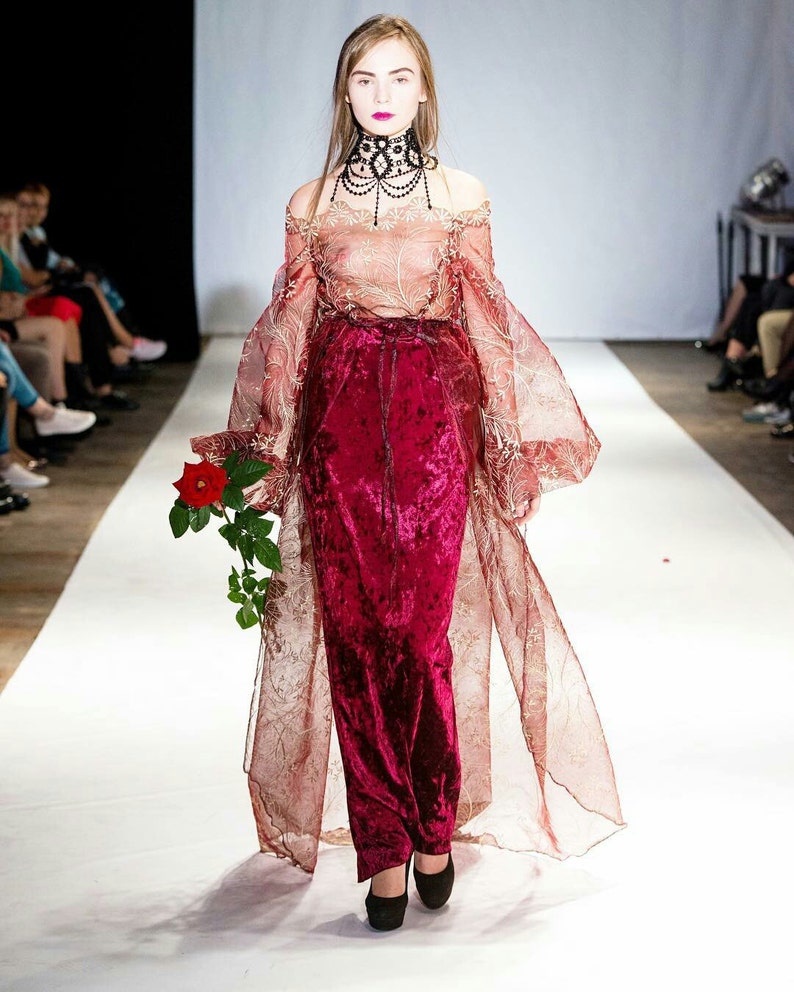 May include: A model walks the runway wearing a long, red velvet skirt with a sheer, gold-patterned overlay. The skirt is paired with a sheer, off-the-shoulder top and a black beaded choker.