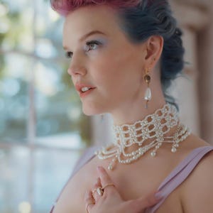 May include: A pearl choker necklace with multiple strands and a delicate, openwork design. The necklace is paired with matching pearl earrings and several gold rings. The model has pink and blue hair and is wearing a lavender top.