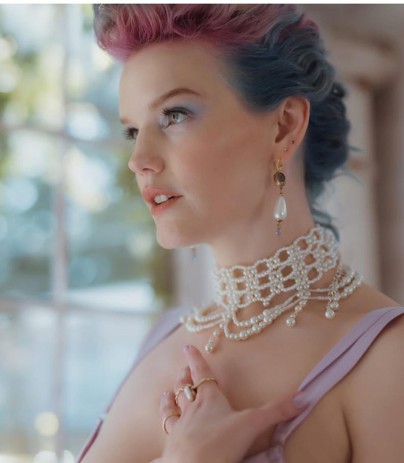 May include: A pearl choker necklace with multiple strands and a delicate, openwork design. The necklace is paired with matching pearl earrings and several gold rings. The model has pink and blue hair and is wearing a lavender top.