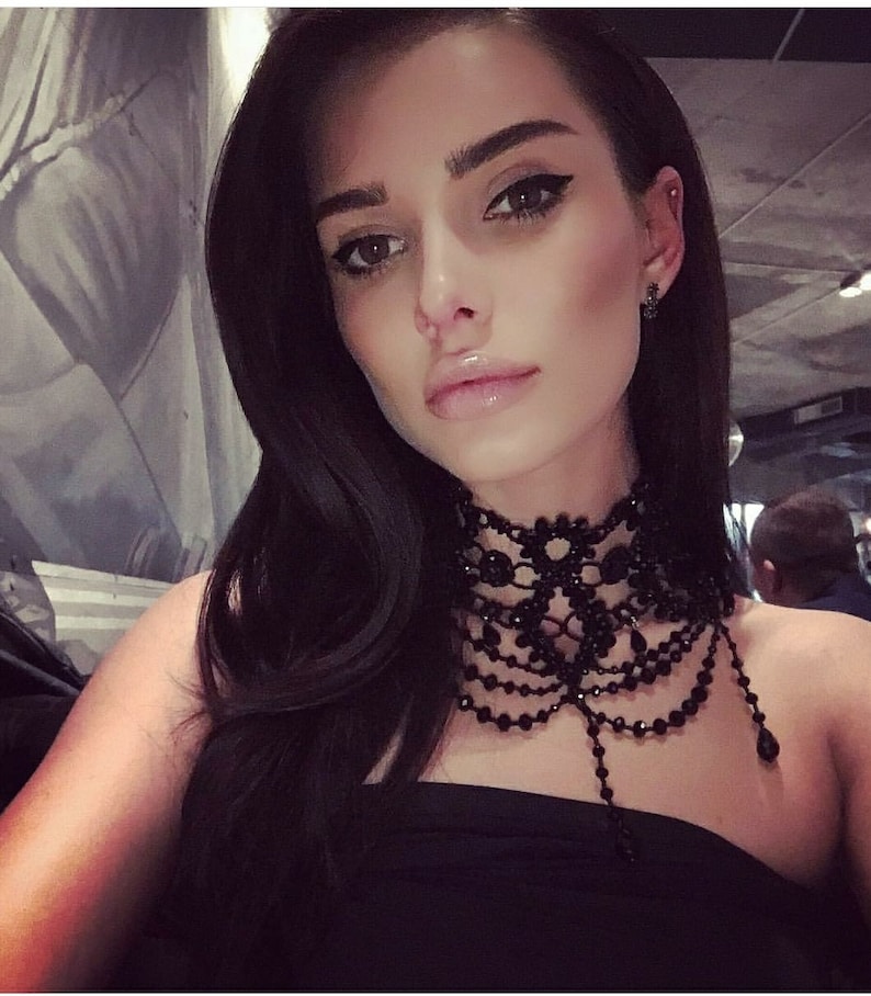 May include: A woman with long black hair wearing a black choker necklace with a beaded fringe. The necklace is made of black lace and beads.