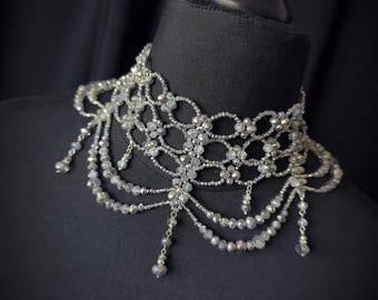 May include: A silver beaded choker necklace with a delicate floral design and dangling beaded strands. The necklace is displayed on a black mannequin head, showcasing its intricate details and craftsmanship. The choker is a statement piece.