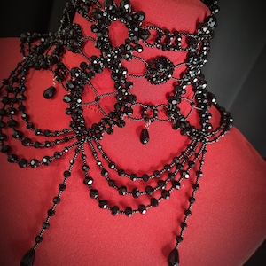 May include: A black beaded choker necklace with multiple layers and dangling beads. The necklace is made of small, black beads that are strung together in a delicate pattern. The necklace is displayed on a red mannequin.
