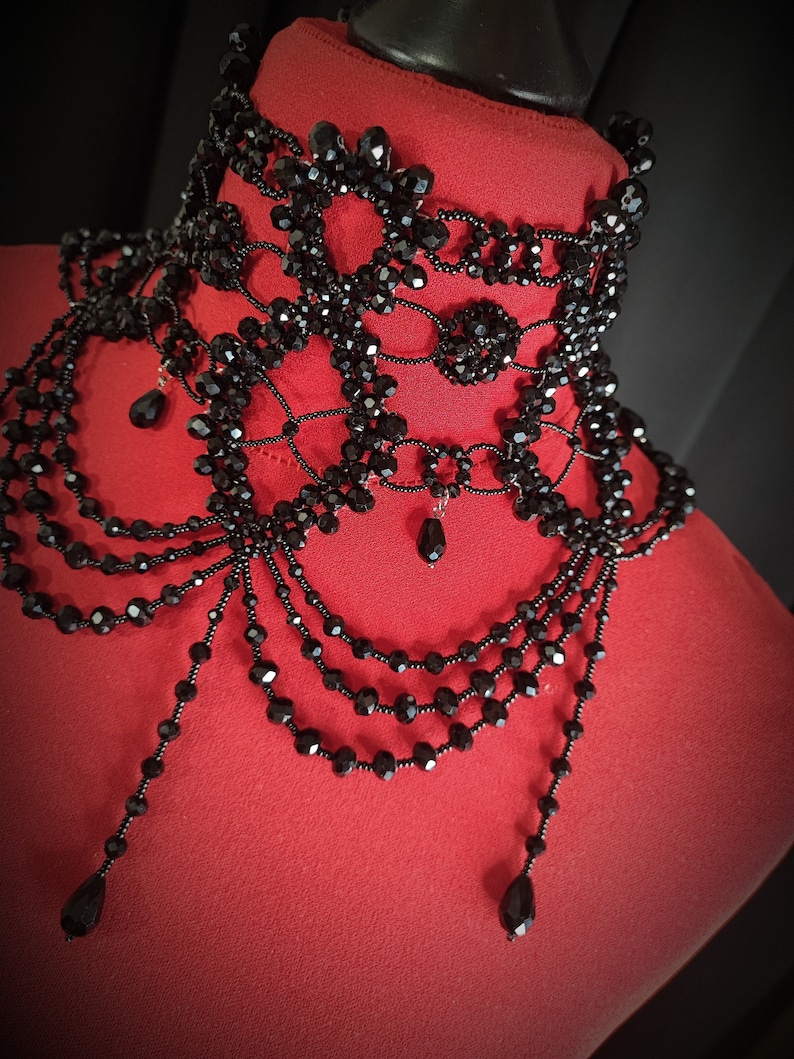 May include: A black beaded choker necklace with multiple layers and dangling beads. The necklace is made of small, black beads that are strung together in a delicate pattern. The necklace is displayed on a red mannequin.