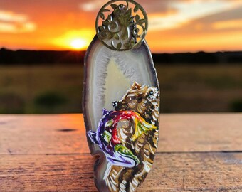 Puede incluir: Un colgante de rebanada de ágata vertical con un oso pardo pintado a mano y un pez colorido de múltiples tonos. La piedra tiene un centro translúcido y un borde exterior oscuro. Una fianza circular de metal dorado con filigrana está unida a la parte superior.