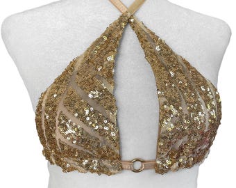 May include: A gold sequin halter top featuring a sheer mesh base with a geometric pattern of metallic gold sequins. The garment has a halter neck strap and a central gold-toned metal ring connecting the two front panels at the lower edge.