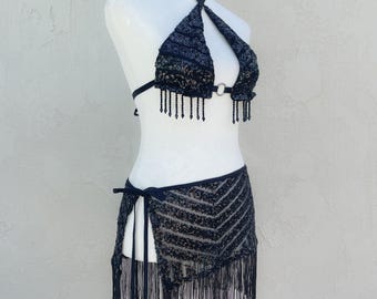 May include: A black two-piece set featuring a halter-style bikini top with sequined chevron patterns, a central metal ring, and dangling beaded fringe. The matching skirt has a side tie closure, a sequined chevron design, and long fringe trim.