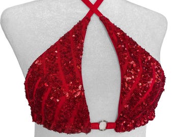 May include: A red halter top featuring a dense covering of shimmering sequins arranged in a wavy, vertical pattern. The garment has a deep, open neckline connected at the centre by a small, circular silver-toned metal ring at the base.