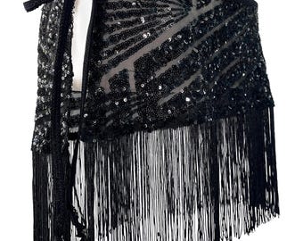 May include: A black wrap skirt featuring a sheer mesh base adorned with dense black sequin patterns in a geometric design. The garment includes a long, flowing black fringe trim along the bottom edge and a black fabric tie closure at the waist.