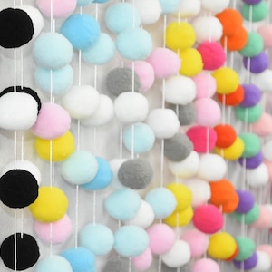 Puede incluir: Una colorida guirnalda de pompones con una variedad de colores, incluyendo negro, blanco, rosa, amarillo, azul, gris, naranja, rojo y morado. Los pompones están ensartados en cuerdas blancas, creando una estética decorativa y juguetona.