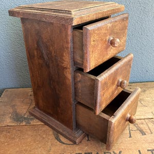Peut inclure: Un petit meuble ancien en bois avec trois tiroirs, chacun avec un bouton rond. Le meuble est de couleur marron chaud et repose sur une surface en bois. Les tiroirs sont partiellement ouverts, révélant l'intérieur.