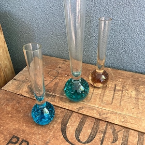 May include: Three clear glass bud vases with rounded, textured bases in blue, amber, and clear. The vases are displayed on a wooden surface with printed text. The vases are of varying heights.