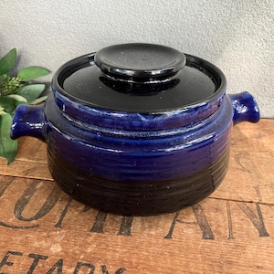 May include: A ceramic pot with a lid, in deep blue and black. The pot has two handles and a circular lid. The pot is sitting on a wooden surface. The pot is approximately 15 cm in diameter.