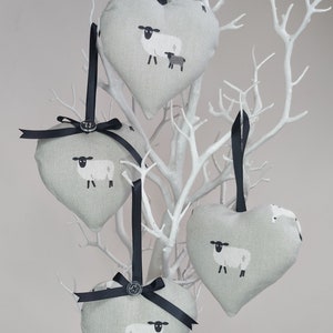 May include: Four grey heart-shaped fabric ornaments with black ribbon hangers and black buttons. Each ornament features a white sheep and a black lamb printed on the front.