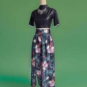 May include: A black faux leather crop top with short sleeves and a high neckline is paired with a high-waisted, floral print skirt. The skirt features a dark background with colorful rose patterns. A silver necklace completes the ensemble.