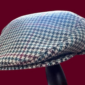 May include: A flat cap featuring a multi-colored houndstooth pattern in shades of green, cream, and dark brown, accented by thin, intersecting red and blue lines that create a subtle plaid effect across the textured fabric of the headwear.