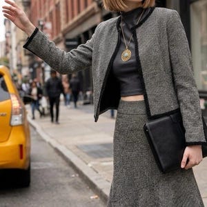 May include: A grey and black tweed jacket with black trim and a matching tweed skirt with a flared hem. The outfit is paired with a dark grey mock neck crop top, a gold pendant necklace with a blue stone, and a black leather clutch bag.