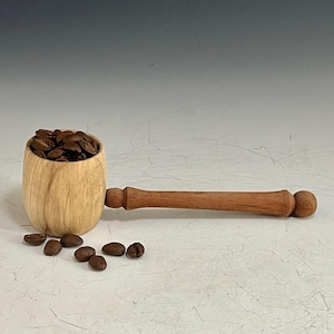 May include: A wooden coffee scoop filled with roasted coffee beans. The scoop has a light wood bowl and a long, reddish-brown handle. Several coffee beans are scattered around the scoop on a textured surface.