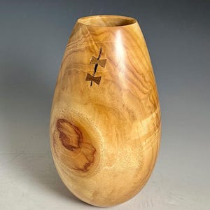 May include: A light brown wooden vase with a smooth, polished finish and a prominent natural wood grain pattern. The vase features a large circular knot on the lower side and two dark, butterfly-shaped wooden inlays securing a vertical crack.