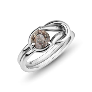 May include: A silver ring with a knot design and a round, grey, salt and pepper diamond.