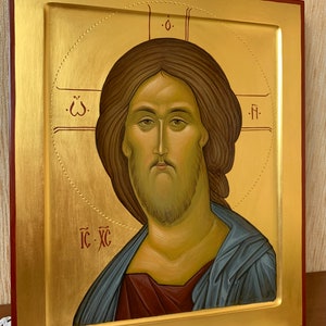 Op de afbeelding: Een religieuze icoon in een lijst met een portret van Jezus Christus. De afbeelding heeft een gouden achtergrond met een halo en een rode rand. De figuur heeft bruin haar en een baard en draagt een blauw gewaad.