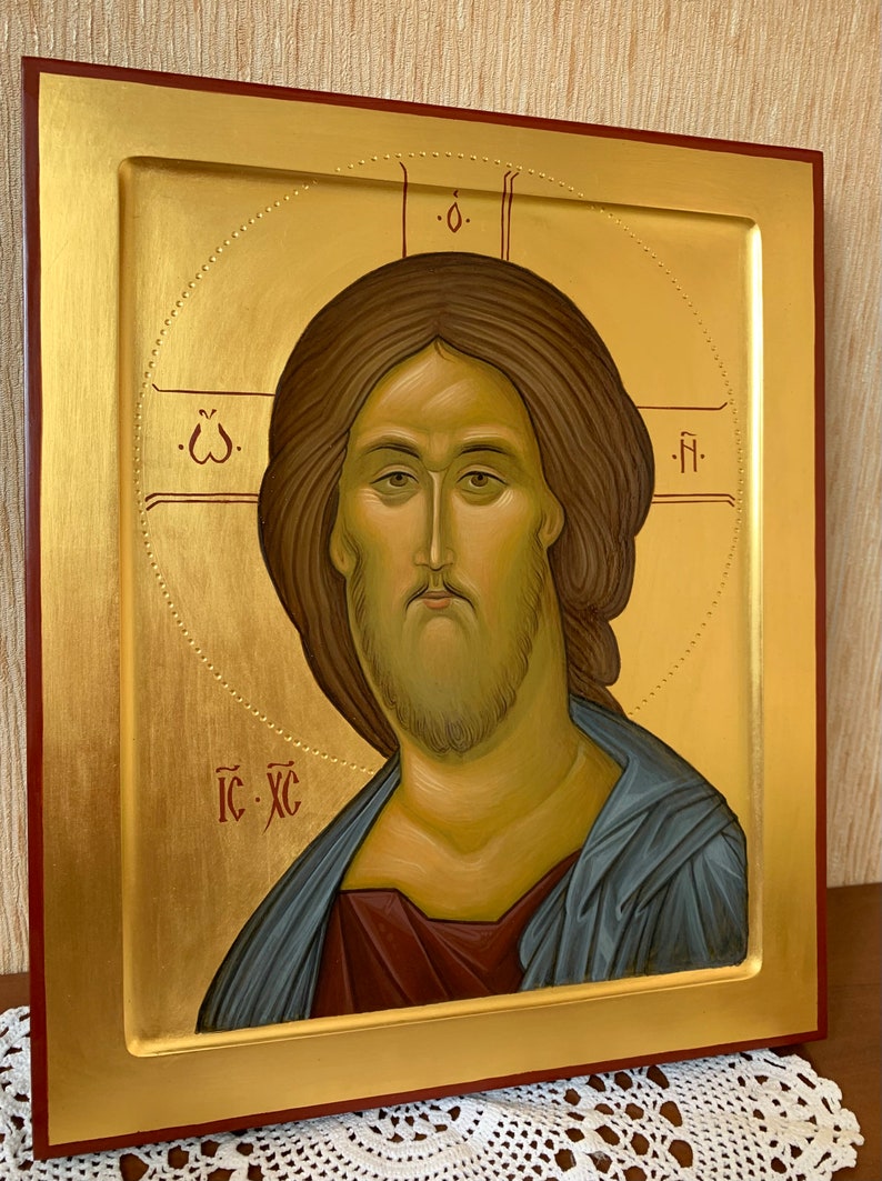 Op de afbeelding: Een religieuze icoon in een lijst met een portret van Jezus Christus. De afbeelding heeft een gouden achtergrond met een halo en een rode rand. De figuur heeft bruin haar en een baard en draagt een blauw gewaad.