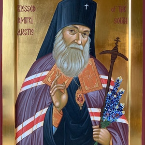 May include: An icon of Blessed Dmitri, Apostle of the South, a religious painting depicting a man with a white beard wearing a black hat and a purple robe. He is holding a staff with a cross on top and a bouquet of blue flowers.