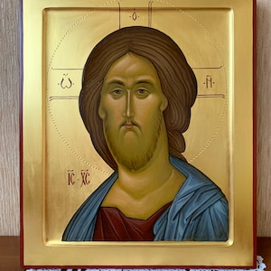 Op de afbeelding: Een religieus icoon met een portret van Jezus Christus. De afbeelding toont Jezus met bruin haar en een baard, gekleed in een blauwe mantel over een rood gewaad. De achtergrond is goudkleurig met rode accenten, omlijst in een houten lijst.