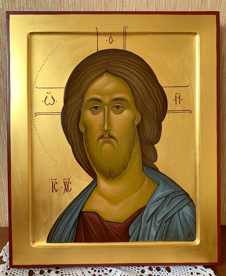 Op de afbeelding: Een religieus icoon met een portret van Jezus Christus. De afbeelding heeft een gouden achtergrond en een gouden lijst met een rode rand. Jezus wordt afgebeeld met bruin haar en een baard, gekleed in een blauw gewaad. Religieuze tekst is in het rood.