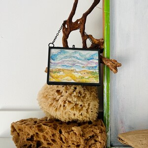 May include: A framed watercolor painting of a landscape with a blue sky, white clouds, and a brown field. The painting is hanging from a driftwood branch and is displayed on top of two natural sea sponges.