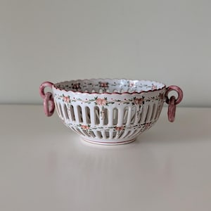 May include: A white ceramic bowl featuring a scalloped rim and vertical cut-out slats. The exterior is decorated with hand-painted pink floral motifs and green foliage. Two pink ring-shaped handles are attached to the sides of the bowl.