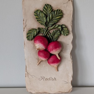 May include: A rectangular wall plaque featuring a 3D relief of radishes. The radishes are red with white tips, and green leaves. The word "Radish" is written in cursive at the bottom. The plaque has a textured, aged appearance.