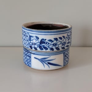 May include: A cylindrical ceramic container featuring a white base with blue hand-painted floral and botanical motifs. The design includes a cross-hatched grid pattern on the sides, with a dark interior and a smooth, glazed finish throughout.