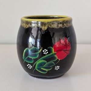 May include: A small, round, black ceramic vase with a yellow and brown rim. The vase is decorated with a red rose, green leaves, and white dots. The vase is approximately 4 inches tall.