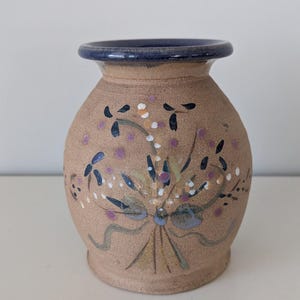 May include: A small, handcrafted ceramic vase with a beige body and a dark blue rim. The vase is decorated with a floral design in shades of purple, blue, white, and green. The vase is approximately 6 inches tall.