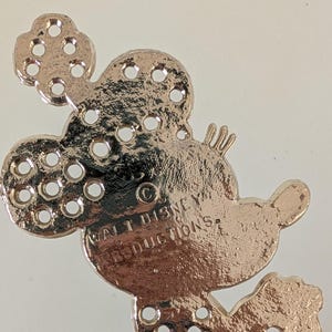 May include: A rose gold-colored Mickey Mouse-shaped charm with multiple small holes. The charm has the text "WALT DISNEY PRODUCTIONS" and a copyright symbol. The charm is likely a decorative element for jewelry or crafts.