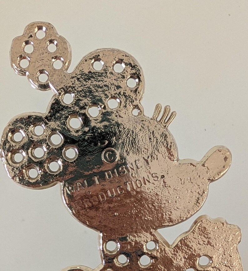 May include: A rose gold-colored Mickey Mouse-shaped charm with multiple small holes. The charm has the text "WALT DISNEY PRODUCTIONS" and a copyright symbol. The charm is likely a decorative element for jewelry or crafts.