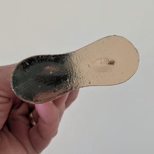May include: A metallic gold-colored bottle stopper with a rounded paddle shape. The stopper has a textured surface and is held by a hand against a white background. The stopper is designed to seal a bottle.