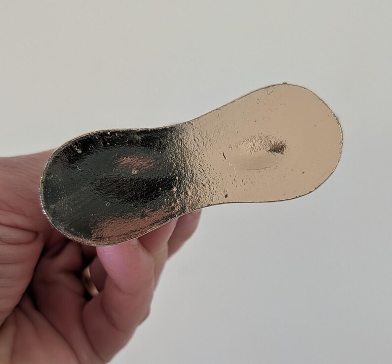 May include: A metallic gold-colored bottle stopper with a rounded paddle shape. The stopper has a textured surface and is held by a hand against a white background. The stopper is designed to seal a bottle.